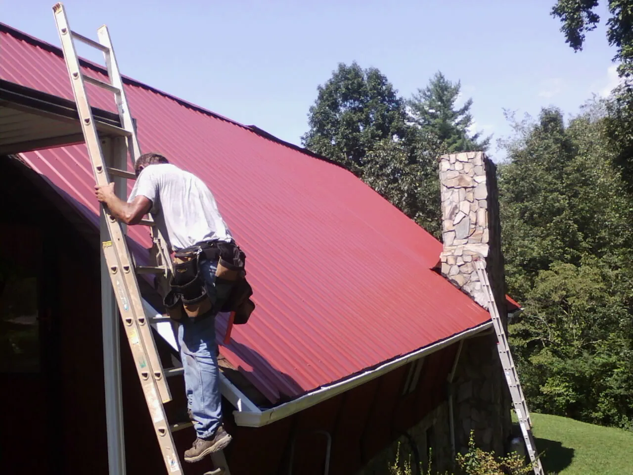 Roofer on a ladder beside a red metal roof and stone chimney — Soffit & Fascia Repair by Roofing Brothers in Boxholm